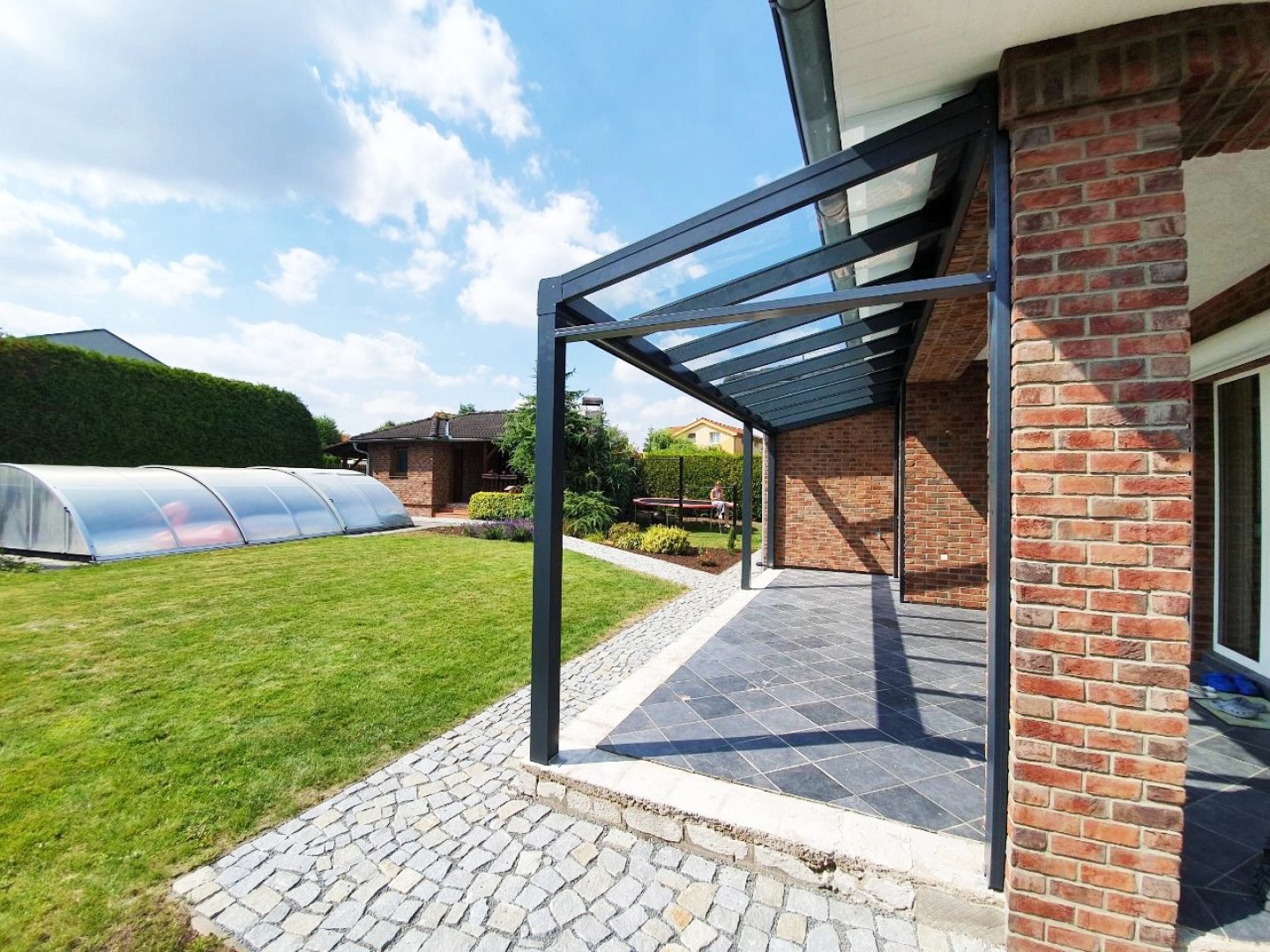 Modern, antracit színű alumínium pergola üvegtetővel egy téglaház mellett, kerttel és medencével.