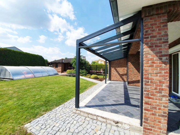 Modern, antracit színű alumínium pergola üvegtetővel egy téglaház mellett, kerttel és medencével.
