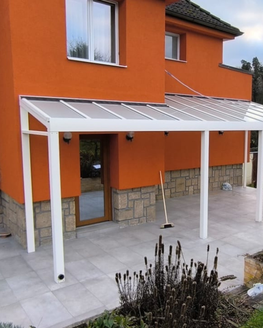 Egy modern, fehér alumínium pergola, amely egy narancssárga, üvegtetős és csempézett házhoz van rögzítve