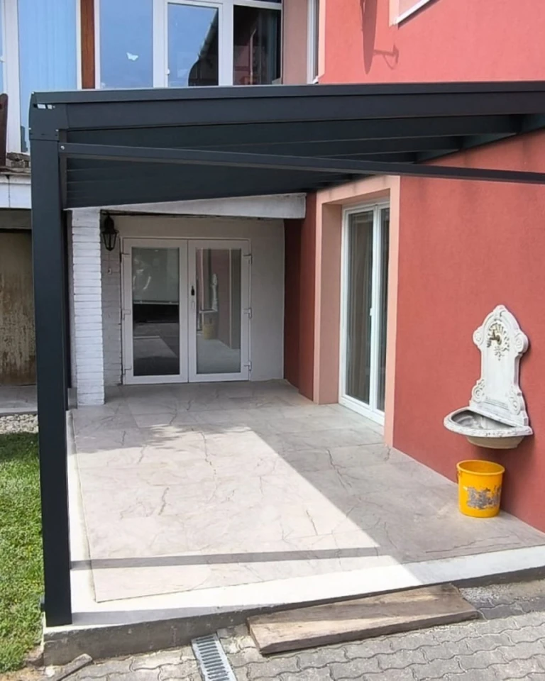 A Robust pergola modern alumínium szerkezete a vörös homlokzatú ház mellett, terasszal.