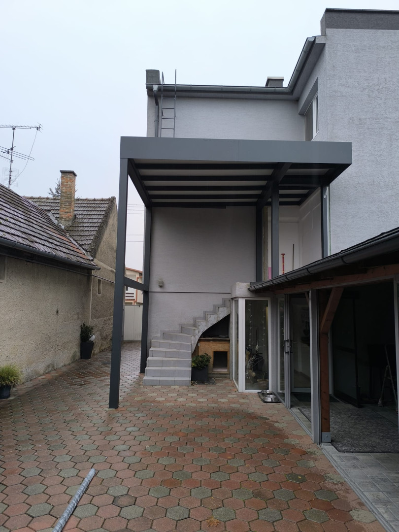 Egy családi ház teraszán, a külső lépcsőház felett felszerelt, egyedi gyártású, modern szürke alumínium pergola.