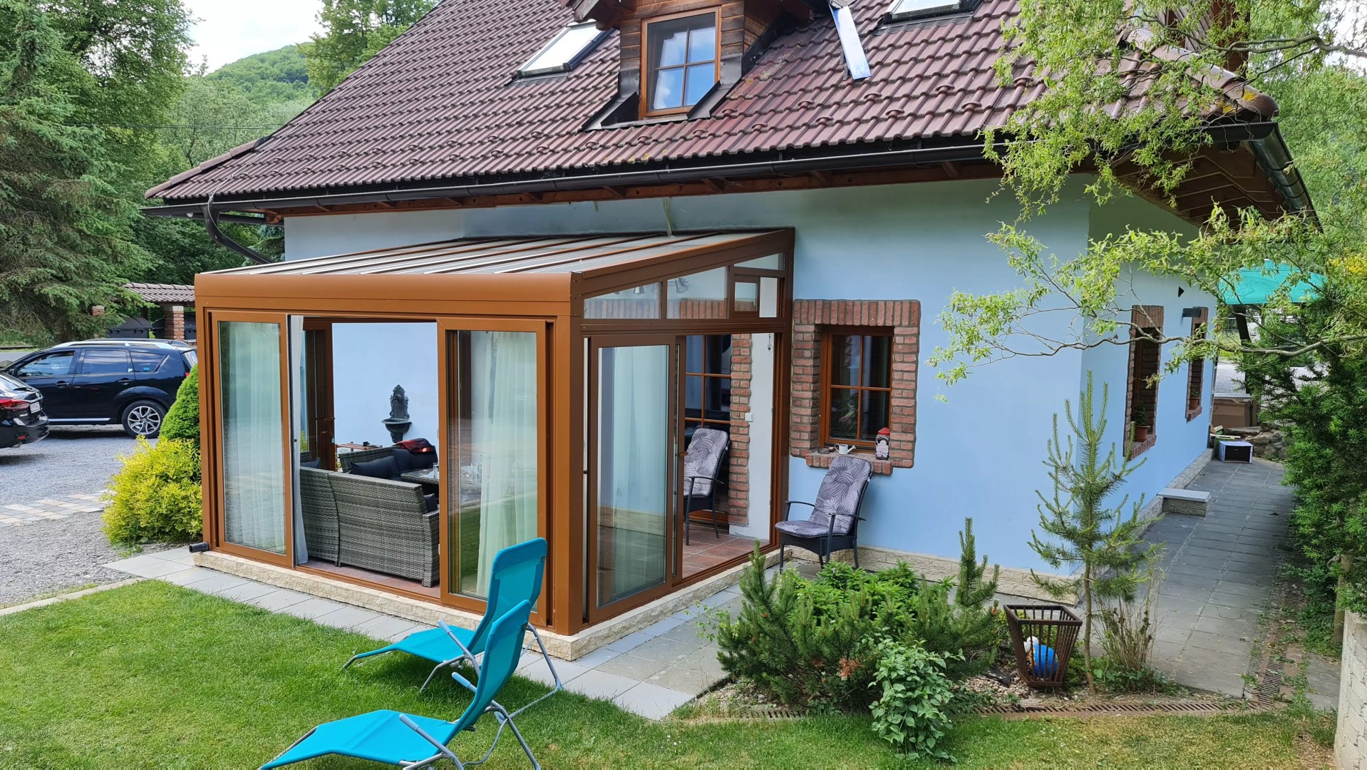 Barna színű, üvegezett alumínium ROBUST pergola, amely egy családi ház mellé épült, kerti bútorokkal.