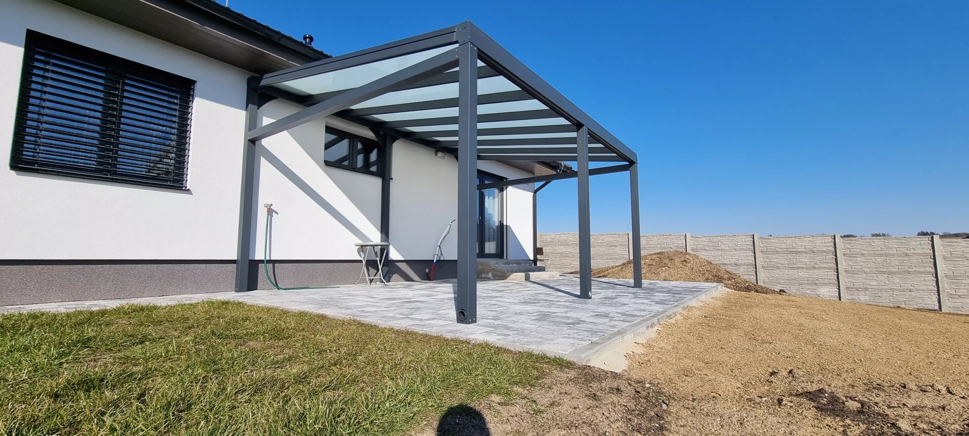 Egy családi házhoz épített, üvegtetős, modern, antracit színű alumínium pergola, amely egy csempézett teraszon áll.