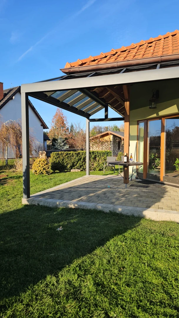 Egy családi ház teraszán álló, modern, szürke alumínium pergola üvegezett tetővel, kerti bútorokkal.