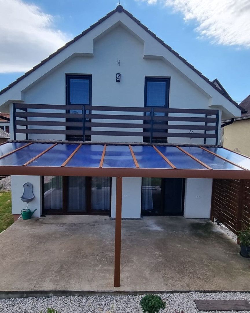 Barna alumínium pergola üvegtetővel, amely egy erkéllyel rendelkező családi házhoz épült.