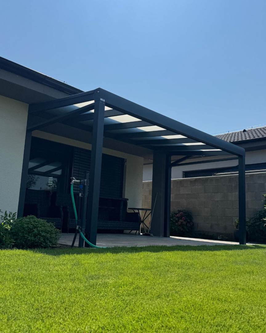 A házhoz csatlakozó, modern, sötét szürke alumínium pergola, üvegtetővel és gondozott pázsittal
