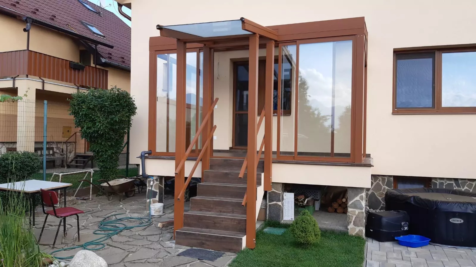 Barna, üvegezett alumínium pergola, amely egy családi ház bejárati lépcsője felett nyújt fedelet.