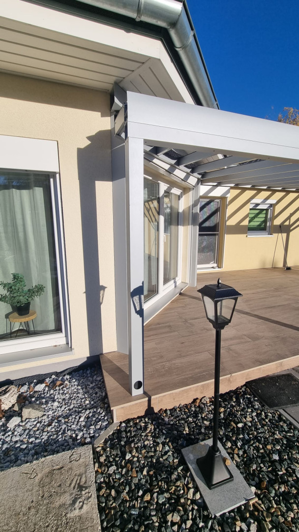 A fehér alumínium pergola rögzítésének részlete a ház homlokzatához, fa díszítésű teraszon.