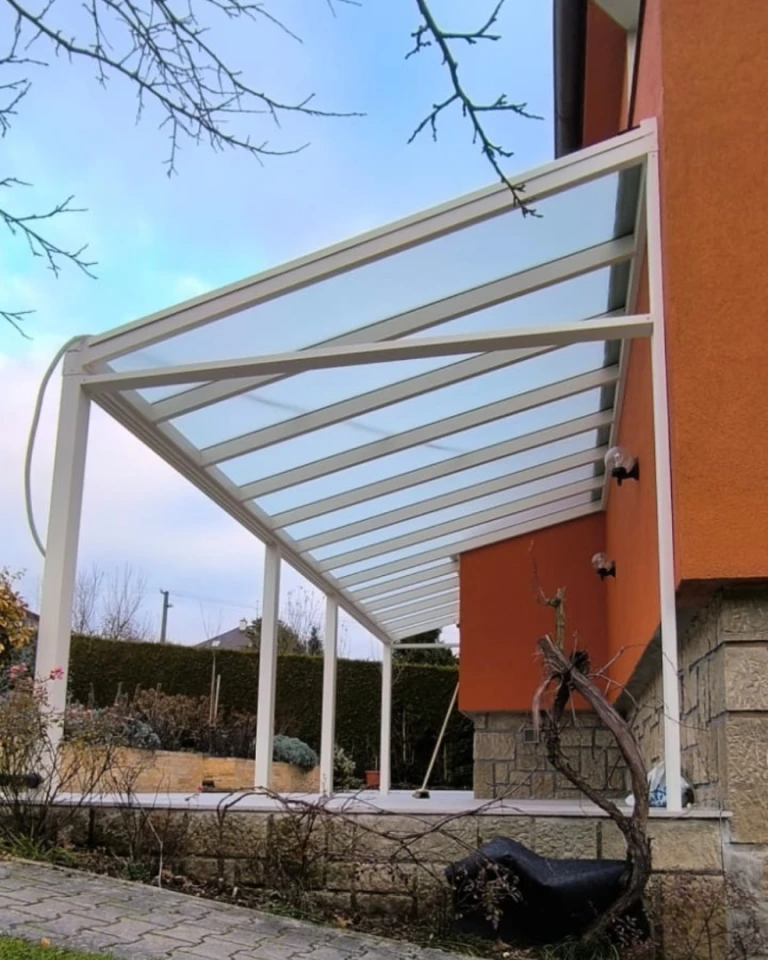 Egy modern, fehér alumínium pergola üvegtetővel, amely egy narancssárga családi házhoz van rögzítve