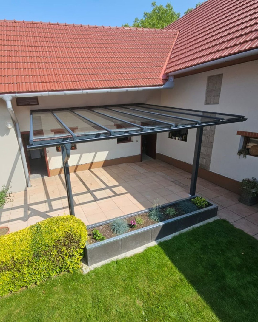 Modern, üvegtetős alumínium pergola, amely a ház falához van rögzítve, burkolt terasszal.