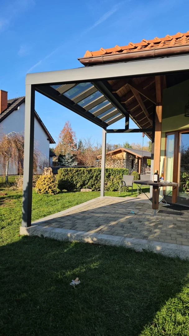 Modern, szürke alumínium pergola üvegezett tetőrészzel egy kőburkolatú teraszon, egy kerttel rendelkező családi ház mellett.