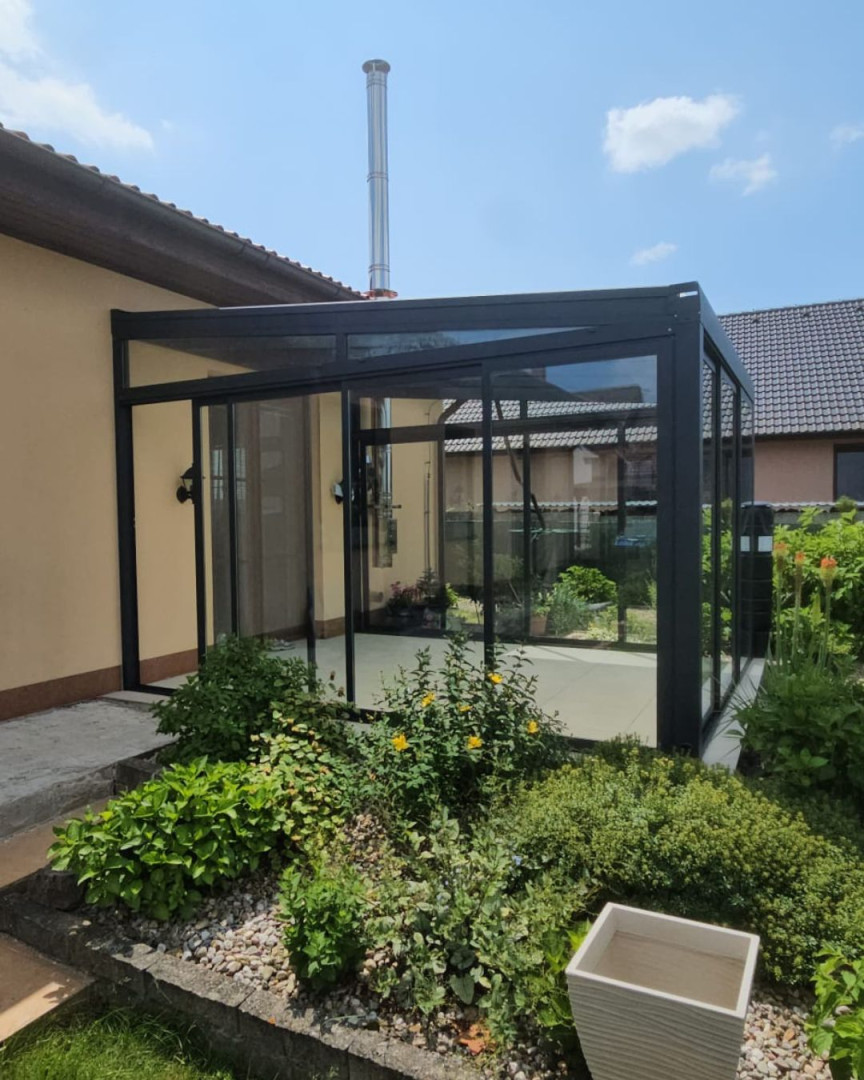 Egy családi házhoz épült modern, üvegezett, fekete keretes, tolóajtós toldalék, amelyet kert vesz körül.