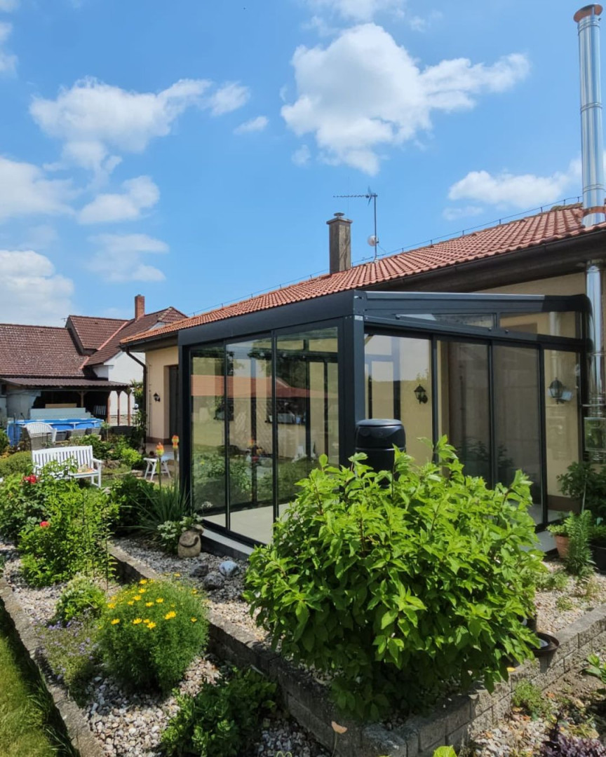 Egy családi ház mellett elhelyezkedő, gondozott kerttel rendelkező, modern, üvegezett télikert antracit színű vázszerkezettel